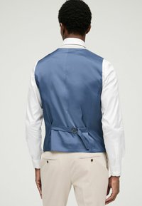 Gilet en satin bleu au design épuré, doté d'une ceinture arrière et d'une boucle ajustable, associé à une chemise blanche à manches longues.