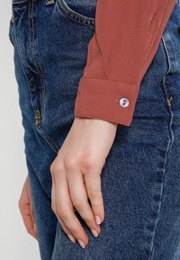 Blouse à manches longues de couleur rouille avec une finition texturée, dotée d'un poignet arrondi et d'une fermeture à un bouton, associée à un jean en denim bleu.