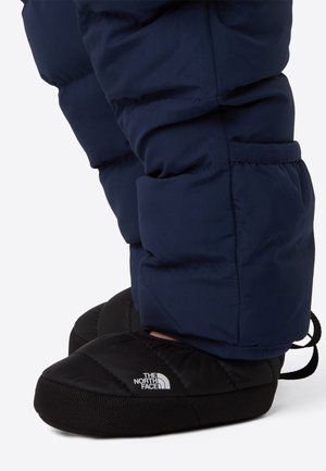 Черни шити пантофи на North Face с плъзгане, носени с тъмносини подплатени панталони, показани от страничен ъгъл на бял фон.