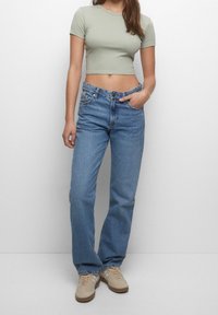 Lysegrøn cropped t-shirt, højttaljede blå jeans med en afslappet pasform, og beige sneakers med hvide accenter, sat mod en ensfarvet baggrund.