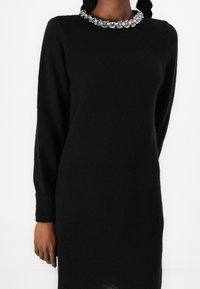 Robe noire en tricot avec manches longues et encolure arrondie ornée de décorations florales. Texture douce et coupe mi-longue.