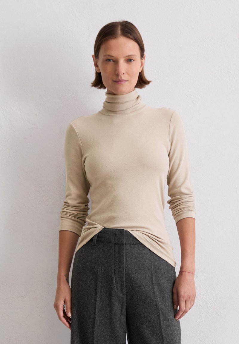 Beige coltrui met lange mouwen, getailleerd ontwerp en een knoopdetail bij de taille, gecombineerd met hoge grijze broek.