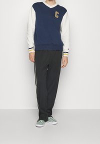 Mikina v námořnické modři a krémové barvě s výstřihem do V a aplikací "C", doplněná černými kalhotami s pruhy po stranách a zelenými kostičkovanými teniskami.