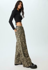 Donna con lunghi capelli scuri, che indossa un top cropped nero a maniche lunghe, pantaloni a gamba larga con stampa leopardata e scarpe con plateau nere, in piedi di lato.