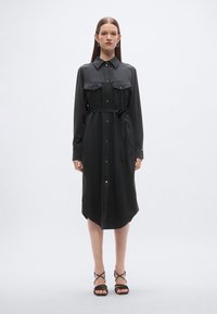 Μαύρο φόρεμα πουκάμισο με μακριά μανίκια, κούμπωμα με κουμπιά, δύο τσέπες στο στήθος, ζώνη στη μέση και καμπυλωτό τελείωμα. Φορεμένο με μαύρα σανδάλια με λουράκια.