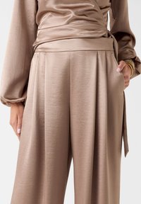 Taupefarbenes Satin-Wickelshirt kombiniert mit hoch taillierter, weit geschnittener Hose, Hand in der Tasche und goldene Armbänder am Handgelenk.