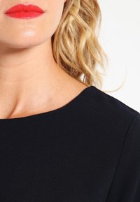 Abito blu navy con una texture liscia, caratterizzato da un collo tondo. Le labbra di un rosso brillante contrastano con il tessuto. I capelli sono ondulati e biondi.