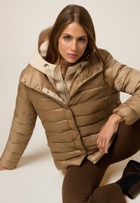 Puffer jacket in light brown with a quilted pattern, featuring a beige hood and zipper. Smooth texture with button accents.