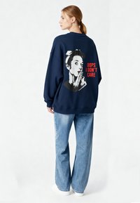 Felpa oversize blu navy con una grafica in bianco e nero sul retro e il testo "OOPS I DON'T CARE" in rosso. Indossata con jeans azzurri.