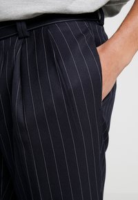 Marinblå randiga byxor i en texturerad tyg. Har en plisserad front och sidfickor, med en midja synlig från en avslappnad position.