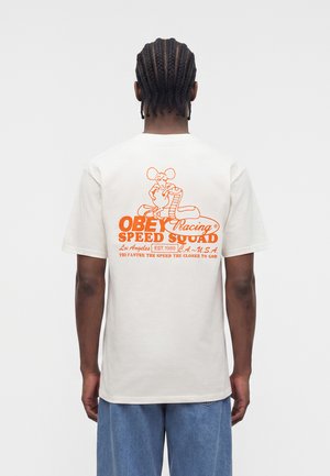 Persona con cabello trenzado que lleva una camiseta blanca con texto naranja "OBEY Racing Speed Squad" y gráfico de un ratón de dibujos animados en la espalda.