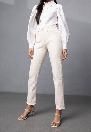 Kobieta ubrana w białą bluzkę z bufkami, kremowe jeansy z wysokim stanem i beżowe sandały na obcasie, stojąca na tle szarej ściany.