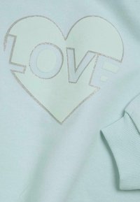 Sweatshirt vert menthe avec un graphique de cœur pailleté portant le mot "AMOUR" au centre. Les poignets côtelés ajoutent de la texture et du détail.