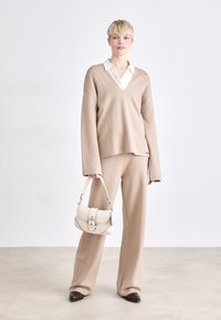 Beige knit sweater with a V-neck and long sleeves, paired with matching pants. A cream handbag with hardware accents is held in one hand.