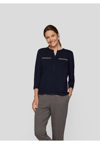 Na্থy-blauer Bluse mit Dreiviertelärmeln und V-Ausschnitt, die zwei vordere Taschen mit dekorativen Akzenten aufweist. Kombiniert mit gemusterten Hosen.