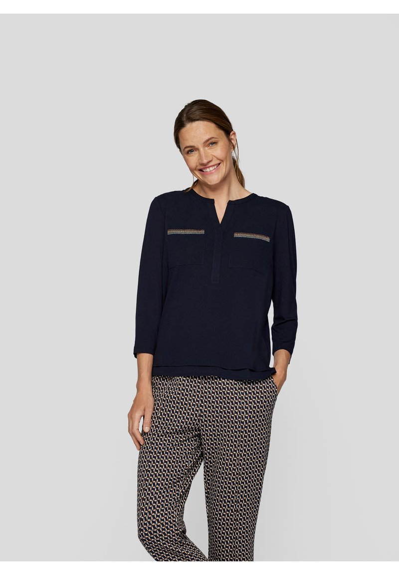 Na্থy-blauer Bluse mit Dreiviertelärmeln und V-Ausschnitt, die zwei vordere Taschen mit dekorativen Akzenten aufweist. Kombiniert mit gemusterten Hosen.