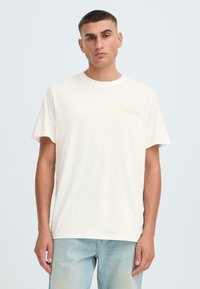 T-shirt en coton couleur crème avec manches courtes, col rond et petit logo jaune "Soulscape" sur le côté gauche de la poitrine. Coupe décontractée.