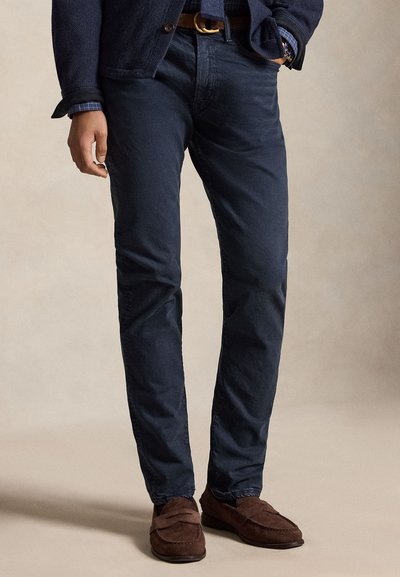 Vyras, apsirengęs tamsiai mėlynomis kelnėmis, rudos spalvos diržu, rudais zomšos loaferiais, mėlyna languota palaidine ir tamsiu tekstūruotu švarku, stovintis su viena ranka kišenėje.