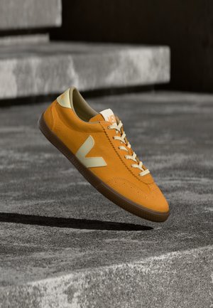 Pantof sport din piele întoarsă portocalie cu accente bej, vârf rotund și talpă de cauciuc texturată. Prezintă șireturi de culoare deschisă și branding pe limbă.