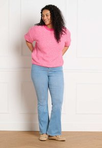 Woman with curly hair wearing a pink striped sweater, light blue jeans, and beige moccasin shoes standing indoors, smiling with hands behind back.