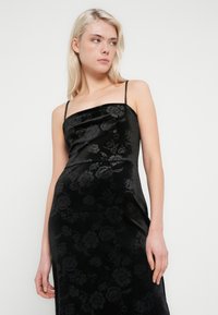 Sort fløyelskjole med blomstertrykk, spaghetti-stropper og strukturert overdel. Glatt tekstur og tettsittende silhuett.