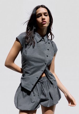 Jeune femme portant une chemise à manches courtes à carreaux boutonnée et un short assorti avec cordon de serrage sur un fond clair uni.