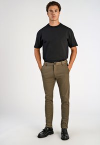 Schwarzes Rundhals-T-Shirt, slim-fit beige Hosen, strukturierter Stoff und schwarze Dressschuhe. Modell steht mit den Händen in den Taschen vor einem neutralen Hintergrund.