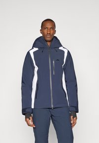 Tengerészkék és fehér síkabát kapucnival, cipz�áras zsebekkel és texturált felülettel rendelkezik. Fekete hardverrel és állítható mandzsettákkal van ellátva.