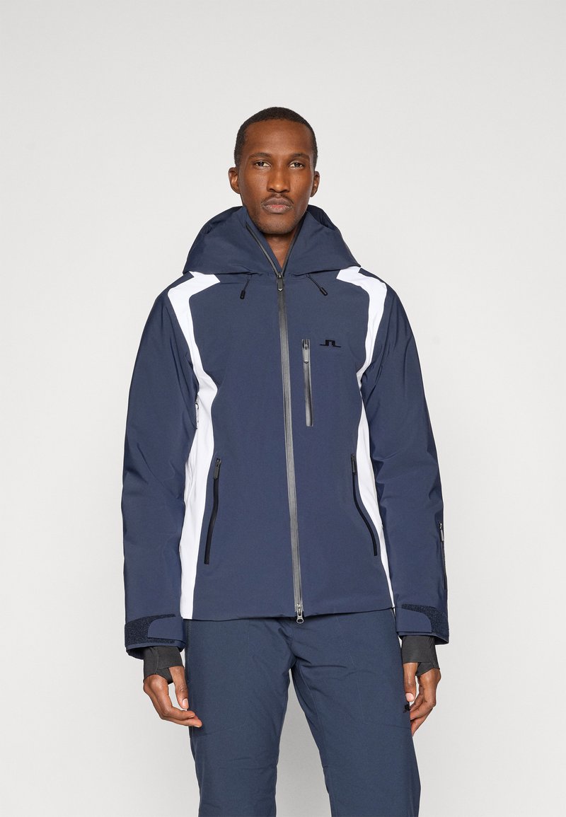 Tengerészkék és fehér síkabát kapucnival, cipzáras zsebekkel és texturált felülettel rendelkezik. Fekete hardverrel és állítható mandzsettákkal van ellátva.