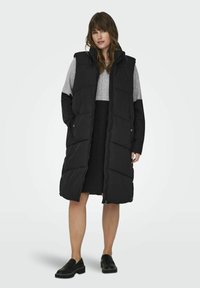 Svart vadderad lång väst med hög krage, blixtlås och två sidofickor. Lager ovanpå en grå topp och svart kjol, ihop med svarta skor.