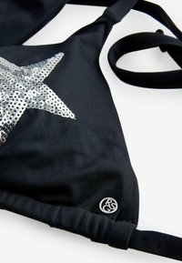Čierny bikiny vrch s veľkým strieborným trblietavým dizajnom hviezdy, nastaviteľnými ramienkami a kovovým logom v tvare kruhu. Mäkka textúra látky.