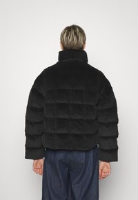 Svart kordflerd pufferjacka med hög krage och quiltat mönster. Har en fyrkantig passform och ribbad textur. Bakvy visas.