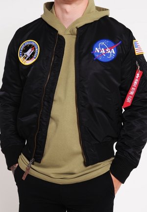 Osoba ubrana w czarną kurtkę lotniczą NASA z naszywkami misji i khaki bluzę z kapturem, ręce w kieszeniach, na jednolitym tle.