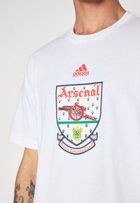 Wit T-shirt met korte mouwen met een kleurrijk Arsenal-logo met een kanon, groene accenten en de tekst 'Victoria Concordia Crescit'.
