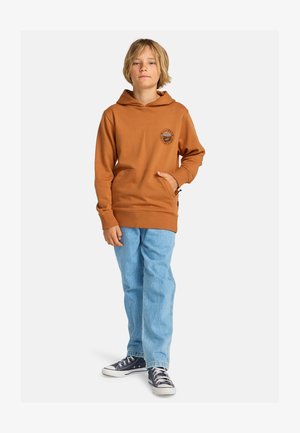 Jongen met blond haar, een bruine hoodie en lichtblauwe spijkerbroek, staand met één hand in de zak en donkerblauwe sneakers op een witte achtergrond.