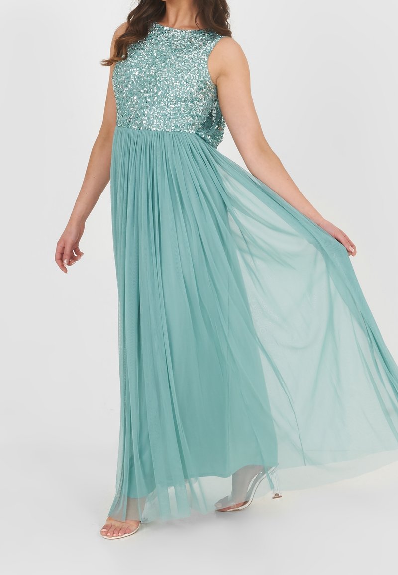 Femme portant une robe de soirée turquoise sans manches avec un corsage à sequins et une jupe fluide en tissu transparent, tenant la jupe d'une main.