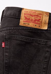Jeans de mezclilla negra con un parche de cuero que lleva gráficos rojos y amarillos, incluyendo "Levi Strauss & Co." y detalles del producto. Etiqueta roja en el bolsillo.