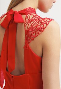Robe rouge avec des détails en dentelle délicate au niveau de l'encolure, un nœud noué à l'arrière et un corsage lisse et ajusté.