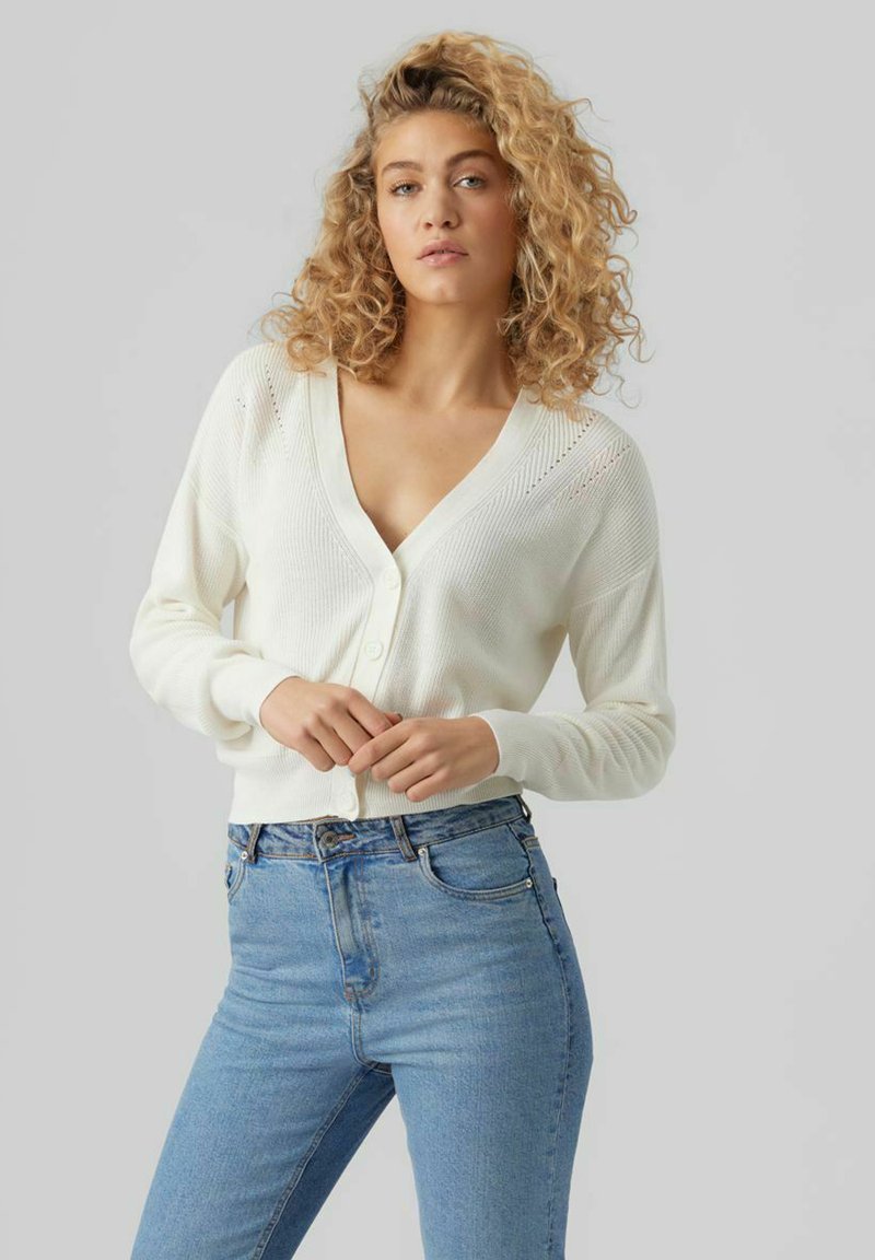 Cardigan en tricot blanc à côtes avec un décolleté en V profond, fermeture à boutons et un léger motif sur les épaules. Associé à un jean bleu taille haute.
