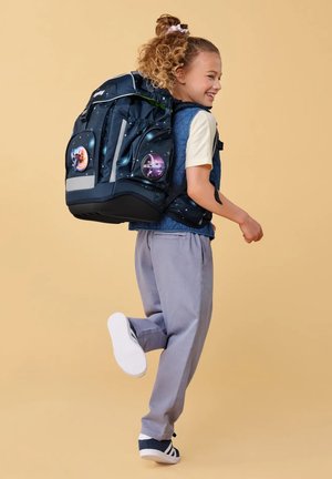 Lächelndes Kind mit lockigem Haar, das einen großen Rucksack mit Weltraummotiv trägt, ein helles Hemd, eine blaue Weste, graue Hosen und Turnschuhe, steht auf einem Bein.