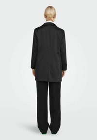 Costume noir comprenant un long blazer avec un col blanc contrastant et un pantalon sur mesure, mettant en valeur une silhouette structurée et une texture lisse.