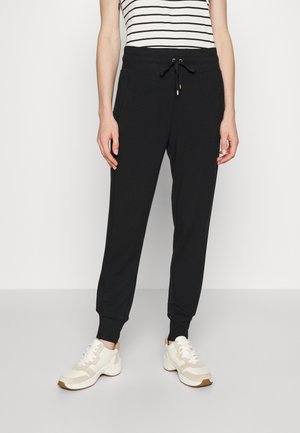 Osoba ubrana w czarne spodnie dresowe ze ściągaczem w pasie, z mankietami na nogawkach, białe i beżowe sneakersy oraz czarno-biały, bezrękawnik w paski.