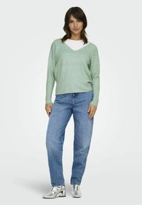 Svetlo zelena v-neck pulover, sproščen kroj, v kombinaciji s svetlo modrimi jeans hlačami in metalno srebrnimi gležnjarji. Mehka tekstura; sproščen dizajn.