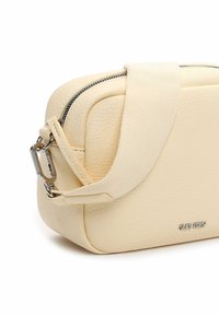 Bolso bandolera de color crema con textura, cremallera plateada, correa de tela ajustable y logo de metal de la marca en la esquina delantera.