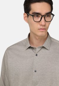 Camicia button-up grigia chiara con tessuto a trama, dotata di colletto alla francese e bottoni scuri. Indossata con occhiali rettangolari neri.