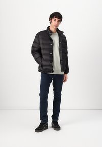 Svart puffjakke med quiltet tekstur og trykknapper, iført over en lys grønn hettegenser, kombinert med mørk denim og svarte joggesko.