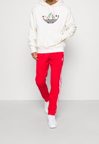 Bílá mikina s kapucí s květinovým motivem a klokáním kapsou, v kombinaci s červenými tepláky s bílými bočními pruhy a logem, doplněná bílými teniskami.