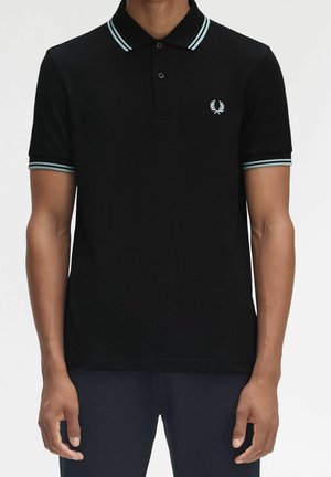 Homme portant un polo noir avec des doubles rayures bleu clair sur le col et les manches, et un petit logo de couronne de laurier sur la poitrine.