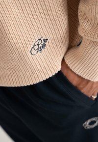 Beige gestrickter Pullover mit besticktem schwarzem Blumenmuster auf der Vorderseite, kombiniert mit schwarzen Hosen und einer Hand in der Tasche.