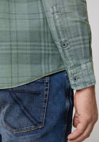Camisa de cuadros verdes con un patrón texturizado, puños abotonados y dobladillo curvado, combinada con jeans de mezclilla azul oscuro que presentan costuras en contraste.